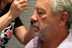 Corte en el pómulo y sangre en el labio, mientras una mano retoca la herida