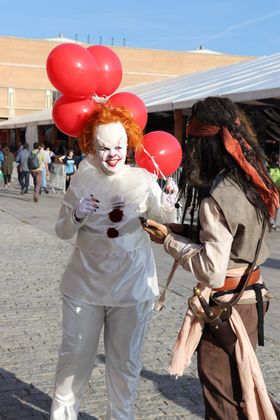 Payaso de traje blanco con un puñado de globos rojos frente a un pirata con pañuelo rojo y espada al cinto, a pleno sol