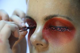 Unos dedos acercan una tira de pestañas postizas negras con puntos rojos a un ojo rodeado de sombra roja; el otro ya la lleva puesta