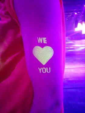 Corazón blanco con las palabras «WE» y «YOU» estarcido en un brazo, sobre un fondo de luces rosas