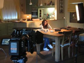 Set de rodaje en una cocina, con una cámara y su monitor en primer plano y una persona sentada a la mesa con un papel