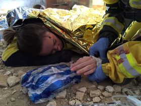 Víctima tumbada entre escombros bajo una manta térmica dorada, con una brecha en la frente
