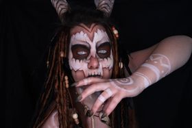 Cara pintada como una calavera blanca y granate, con cuernos, rastas con cuentas de madera y espirales blancas en la mano