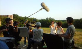 Equipo de rodaje en un campo al atardecer, con una pértiga de sonido, una cámara al hombro y una claqueta
