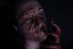 Rostro en penumbra con la piel levantada en ampollas y quemaduras, y una mano ensangrentada junto a la boca