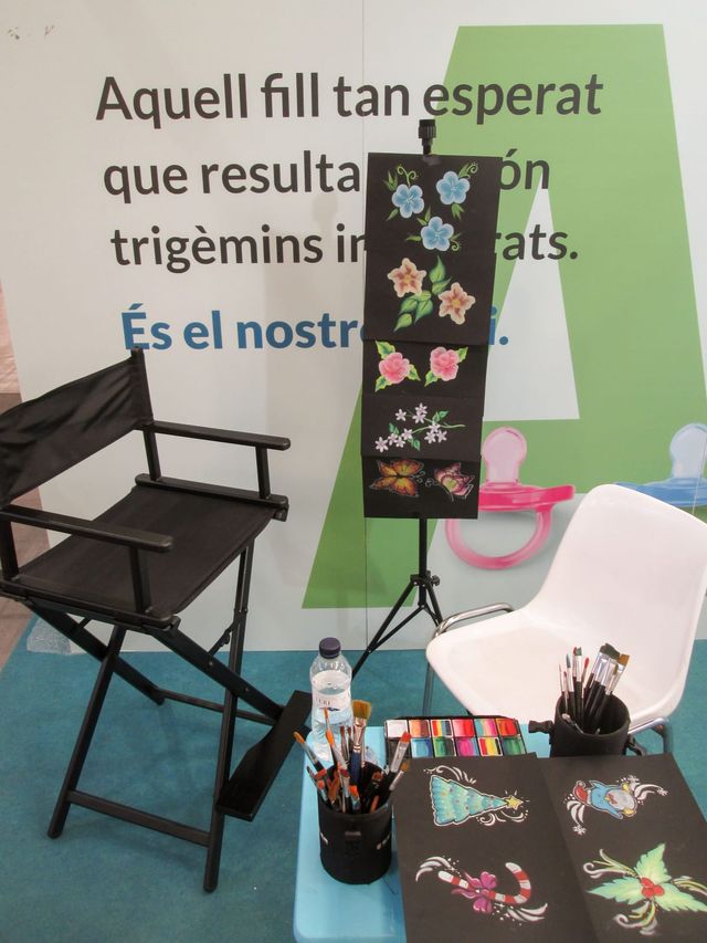 Puesto de maquillaje montado: silla de director negra, cubiletes de pinceles, una paleta de colores y muestras de dibujos navideños y de flores sobre cartulinas negras