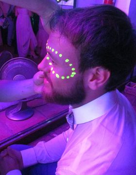 Hilera de puntos amarillo flúor pintada alrededor del ojo bajo luz violeta, mientras una mano sujeta la cara