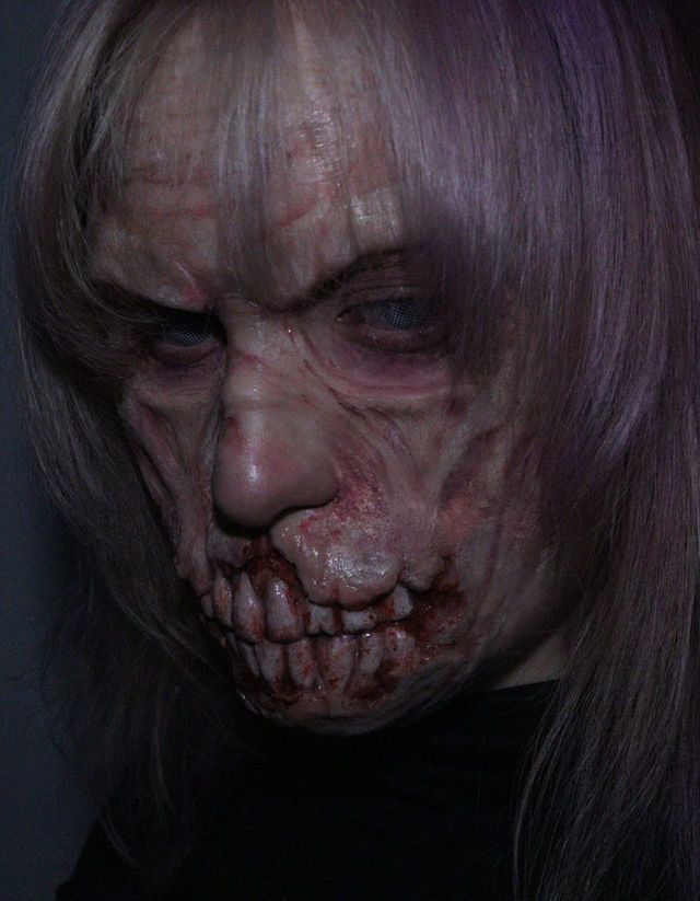 Cara envejecida y podrida, con la boca deshecha en dientes ensangrentados y una melena lila que le cae sobre los ojos