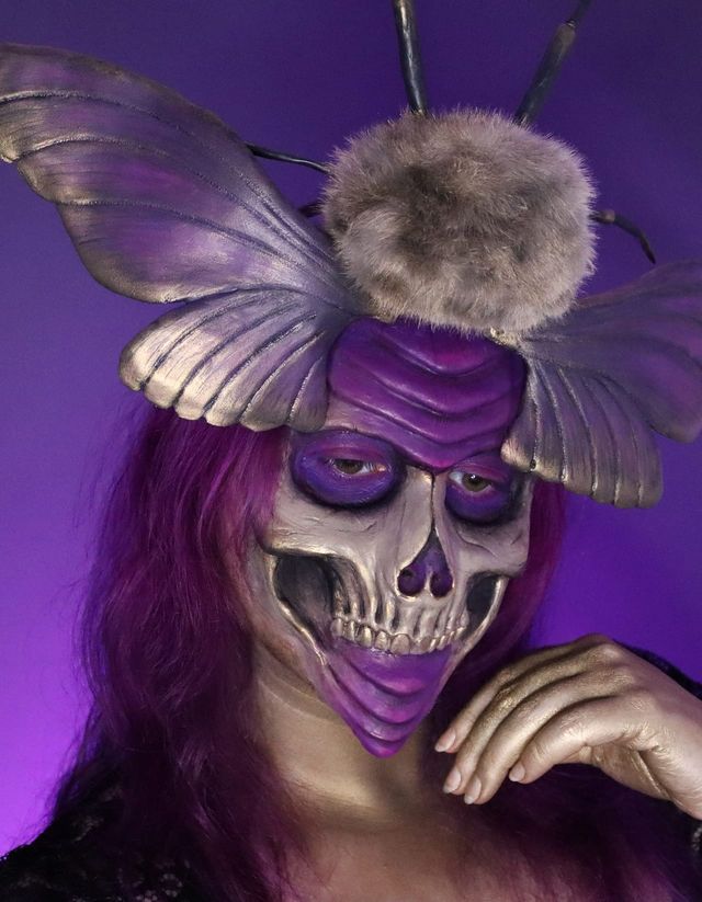 Calavera violeta y dorada pintada sobre la cara, con una gran polilla de alas plateadas posada en la cabeza