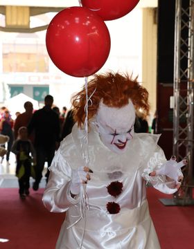 Payaso con la cara pintada de blanco, líneas rojas sobre los ojos y sonrisa roja; traje blanco de raso con pompones rojos y globos rojos, en un recinto lleno de gente