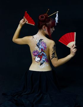 De espaldas y sentada sobre una falda negra: una carpa y flores pintadas en la espalda, caracteres japoneses en el brazo y dos abanicos rojos